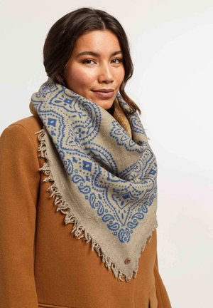 Beige sjaal met blauwe geometrische patronen en franje, om de nek gedrapeerd, gedragen over een bruine jas. Zachte textuur en grote maat.