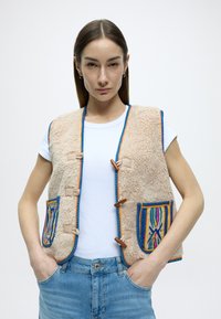 Μπεζ χνουδωτό γιλέκο με μπλε, πορτοκαλί και ροζ υφαντά διακοσμητικά, με δύο μπροστινές τσέπες και ξύλινα κουμπιά τύπου toggle. Φορεμένο πάνω από λευκό μπλουζάκι.