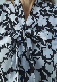 Schwarz-weiße Bluse mit floralem Muster und einer Schleifendetail am Kragen, die eine glatte Textur und ein geschichtetes Blumenmuster vor einem hellen Hintergrund aufweist.