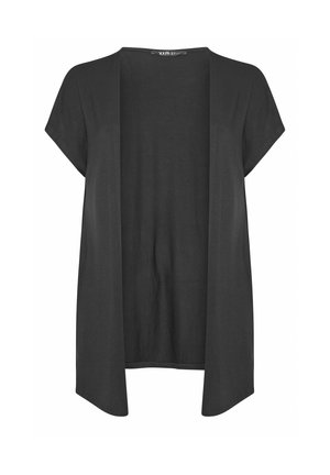 Lyshåret kvinde iført en hvid top, lyseblå højtaljede denimshorts og en ærmeløs sort lang vest mod en ensfarvet baggrund.