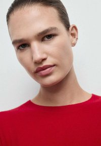 Czerwony sweter ze żebrowanym wzorem i okrągłym dekoltem, zbliżenie na gładką skórę i delikatną kolczyk. Tło w neutralnych kolorach.