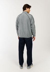 Grau Fleecejacke mit hohem Kragen und schwarzen Paspelakzenten, kombiniert mit dunkelblauen Jeans und gemusterten Sneakers, von hinten betrachtet.