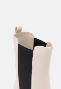 Bottine en cuir blanc et tissu noir, avec une texture lisse, un design fendu et une boucle au talon pour un enfilage facile.