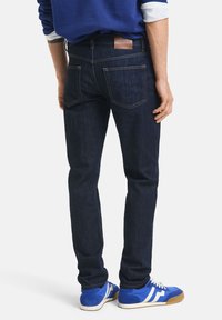 Dark blue denim jeans with a slim fit, featuring back pockets, contrasting stitching, and a brown leather brand patch at the waist.