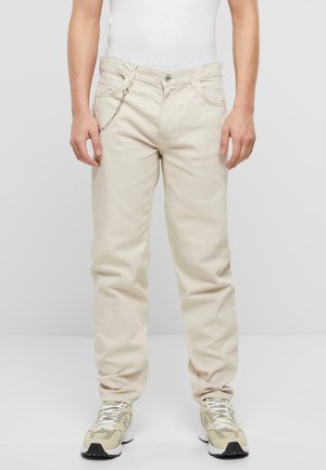 Pantalones de algodón beige con corte recto, que presentan bolsillos frontales, un detalle de cadena en la cintura y combinados con zapatillas de colores claros.