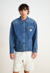 Denim jaka vidējā zilā krāsā ar rāvējslēdzēja priekšpusi, apkakles, divām sānu kabatām un krūšu kabatu, kas rotāta ar kvadrātveida logo plāksteri.