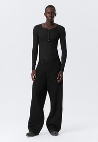 Crna dugosleeve henley majica s dugim rukavima i otvorom za gumbe, uparena s prevelikim crnim trapericama. Model stoji nasuprot svijetle sive pozadine.