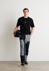 Melna īsām piedurknēm T-kreklā ar nelielu logo, kombinēta ar aplikāciju džinsiem zilo un pelēkā toņos. Modelis tur melnu somu, stāvot pret gaismas fonu.