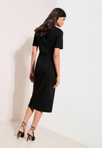 Schwarzes figurbetontes Midikleid mit kurzen Ärmeln und glatter Textur. Kombiniert mit schwarzen, riemchenbesetzten High-Heels, die eine schlanke Silhouette zeigen.