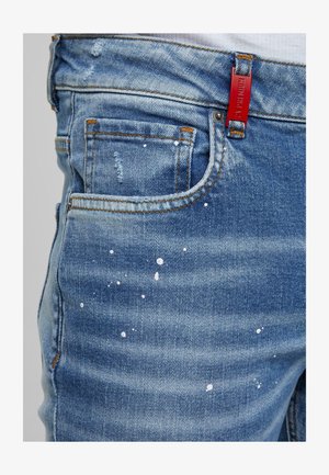 Jeans de mezclilla en azul medio con desgastes, rotos y salpicaduras de pintura blanca; presenta una etiqueta roja en la cinturilla.