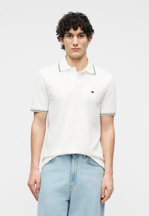 Homme aux cheveux bouclés foncés, portant un polo blanc avec des bordures noires et un jean bleu clair, debout devant un fond uni.