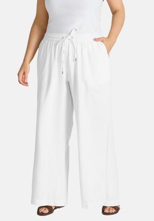 Hvide bukser med vide ben, elastisk talje og snøre, båret med brune sandaler og en hvid ærmeløs top, hænderne i lommerne.