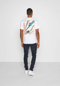 Vit bomullst-shirt med grafiskt tryck på baksidan, med abstrakta former i grönt, orange och blått. Kombinerad med mörka jeans och vita sneakers.