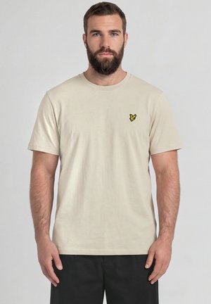 Homme barbu portant un t-shirt beige avec un petit logo d'oiseau jaune sur la poitrine et un pantalon noir, debout devant un fond uni.