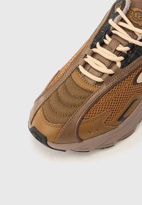 Καφέ αθλητικό παπούτσι με υλικά από δίχτυ και δέρμα, υφή με λεπτομέρειες, μπεζ κορδ όνια και λαστιχένια σόλα με μαύρες λεπτομέρειες.