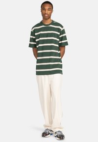 Camiseta a rayas verdes y beige con mangas cortas y cuello redondo, combinada con pantalones sueltos crema y zapatillas deportivas.