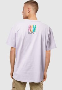 Vaaleanlila puuvillapaita, jossa on värikkäät ja leikkisät tekstit "PRIDE SCOOPS" selässä. Normaali istuvuus, lyhyet hihat ja pyöreä kaula-aukko.