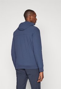 Sweat à capuche bleu marine avec une surface texturée et des bords côtelés. Dispose d'une capuche ajustable et d'un design sans couture au niveau du dos.