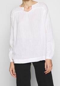 Blouse blanche à manches longues en tissu léger avec un col en V et une texture subtile. Coupe ample, associée à un pantalon noir.