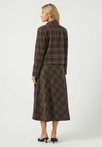 Bruine geruite jas met een rechte snit, gecombineerd met een lange geruite rok. Glad stof met groene en lichtbruine accenten. Zwarte muiltjes met hak.