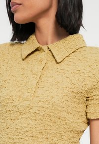 Κοντινό πλάνο μιας γυναίκας που φοράει κίτρινη υφασμάτινη κοντομάνικη πόλο μπλούζα με γιακά και τρία κουμπιά, με το κεφάλι στραμμένο προς το πλάι.