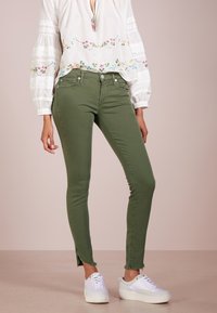 Jeans ajustados verdes con dobladillo deshilachado, material de algodón, bolsillos frontales, y zapatillas blancas a juego con una superficie texturizada. Blusa blanca lisa con bordado.