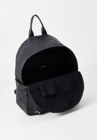 Mochila de cuero negro con forma redondeada, cierre de cremallera y solapa frontal. El interior es de tela negra con una pequeña etiqueta. Cuenta con dos asas superiores.