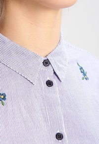 Pruhovaná košile s knoflíky v modré a bílé barvě, s černými knoflíky a modrou květinovou výšivkou na levé straně.