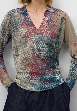 Femme portant un chemisier manches longues en V avec un motif abstrait multicolore à pois et un pantalon taille haute bleu foncé.