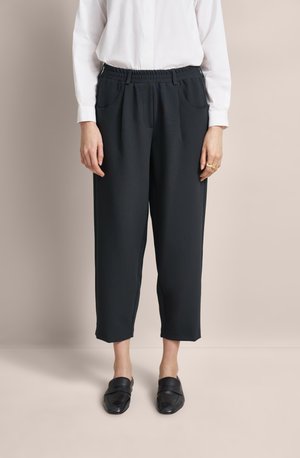Schwarze tapered Hose mit einem elastischen Bund, Seitentaschen und einer glatten Textur, kombiniert mit flachen schwarzen Slippers.