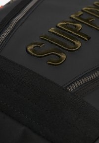 Schwarzer Stoffrucksack mit gesticktem goldenen Text, robusten Reißverschlüssen und strukturierten Trägern, der ein langlebiges und funktionales Design unterstreicht.
