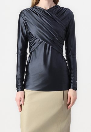 Mujer con una blusa negra ajustada de manga larga con detalle fruncido de envoltura en el pecho, combinada con una falda beige.