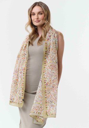 Frau mit welligem blondem Haar, die ein ärmelloses beiges Kleid und einen langen, hellfarbigen Schal mit einem floralen Paisley-Muster trägt.