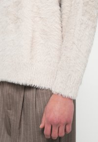 Pulover bej deschis, puțin pufoș, cu finisaj texturat, mâneci lungi și croială lejeră, asortat cu pantaloni maro cu dungi subțiri.