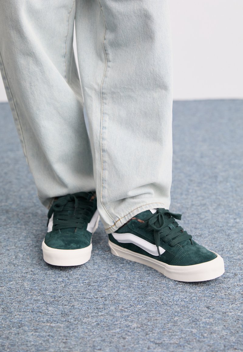 Baskets en suede vert avec des rayures blanches sur les côtés, des lacets plats et une semelle blanche. Associées à un pantalon en denim clair, taille ample.
