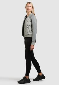 Žena v šedé prošívané bundě, černých legínách a černých teniskách stojí s hlavou mírně otočenou doleva.