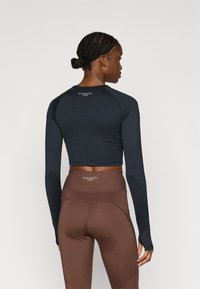 Ilgas rankovėmis trumpintas viršus iš tamsiai mėlynos spalvos audinio, derinamas su aukštos liemens rudos spalvos tamprėmis. Abu drabužiai turi logotipą ant nugaros, pabrėžiantį sportinį dizainą.