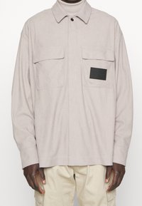 Camicia a quadri beige chiaro con maniche lunghe, colletto, due tasche sul petto e una toppa con logo nero sulla tasca sinistra. Tessuto morbido.