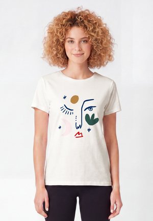Femme aux cheveux bouclés portant un t-shirt blanc avec un motif abstrait de visage en formes bleues, vertes, rouges, marron et roses, debout sur un fond uni.