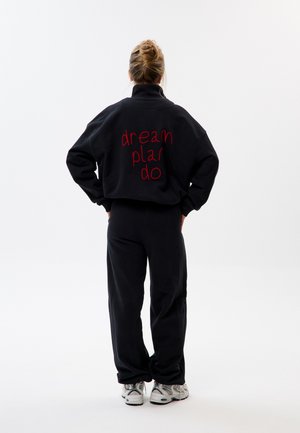 Frau mit blondem Haar zu einem Dutt gebunden, trägt schwarzen Trainingsanzug mit rotem Schriftzug „dream plan do“ auf dem Rücken, steht mit Händen in der Hüfte, weiße Turnschuhe sichtbar.