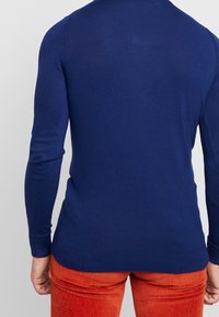 Man som bär en blå långärmad tröja och röda manchesterbyxor, sedd bakifrån mot en enkel bakgrund.