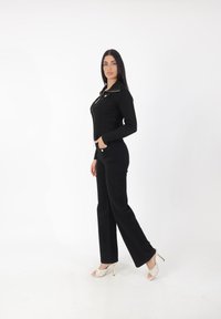 Schwarze gerippte Zip-Jacke mit spitzem Kragen, kombiniert mit eng anliegenden schwarzen Hosen und offenen weißen High Heels mit strukturiertem Muster.