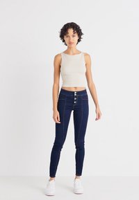 Sötétkék, magas derékú skinny farmernadrág, nagy gombos részletekkel és kontrasztos varrással. Bézs, testhezálló ujjatlan felsővel párosítva.