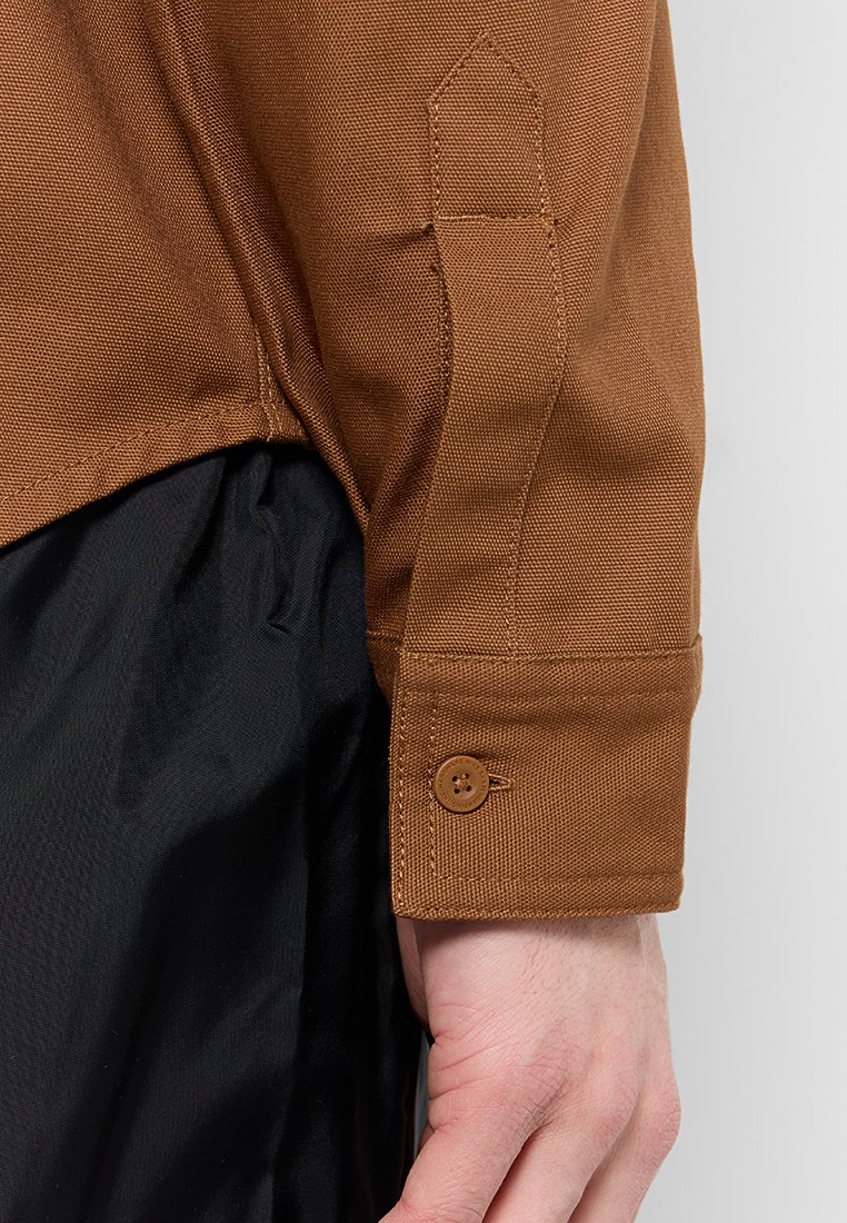 Chemise en tissu marron avec une surface texturée et un poignet boutonné, superposée à un matériau noir. Le poignet dispose d'un bouton et d'un détail de couture en angle.