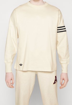 Beige langærmet skjorte med sort Adidas trefoil-logo i kanten og tre sorte striber på højre ærme, parret med beige bukser med maroon "A".