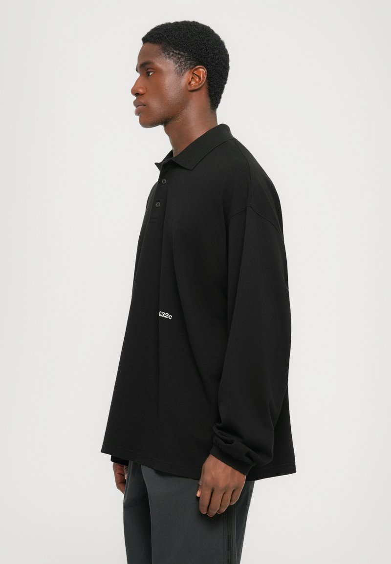 Uomo in polo nera a maniche lunghe e pantaloni scuri in piedi di lato contro uno sfondo bianco semplice.