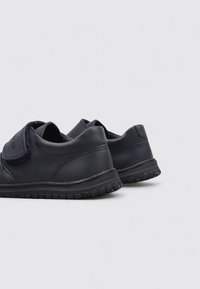 Chaussures pour enfants en cuir noir avec une sangle auto-agrippante et des semelles en caoutchouc texturé, vues de l'arrière sur un fond uni.