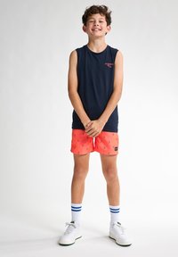 Granatowa koszulka bez rękawów, jasnopomarańczowe spodenki kąpielowe z małymi granatowymi wzorami, białe sneakersy z zielonymi akcentami oraz białe skarpetki w paski.