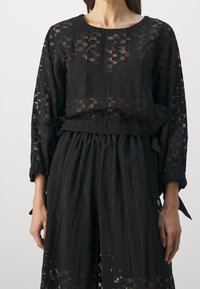 Mustahiili musta paito, jossa on pitkät hihat ja teksturoitu kuvio, tiukka vyötärö ja solmittavat yksityiskohdat hihoissa. Kevyt kangas luo kerroksellisen ilmeen.