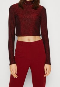 Roter glitzernder, kürzerer Langarm-Top mit hohem Kragen, kombiniert mit maßgeschneiderten roten Hosen. Beide Kleidungsstücke haben eine glatte Textur und eine elegante Passform.